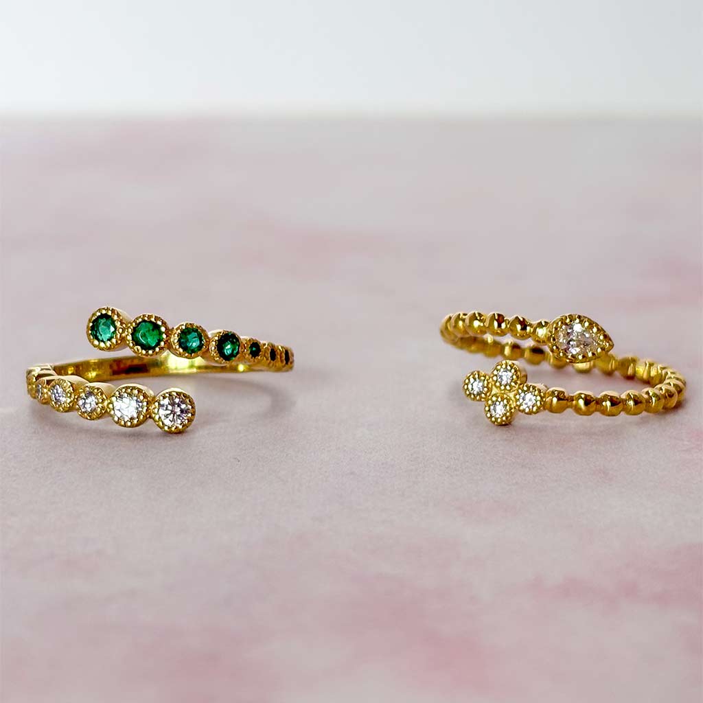 Two gold rings displayed on a soft pink surface. One ring features a row of round green emerald-cut stones, and the other features a row of round clear-cut stones with a small floral cluster at the end.